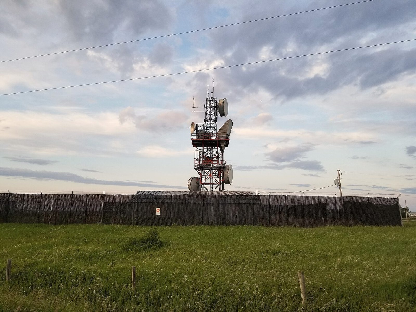 A communications tower standing in an open green field under scattered cloud.